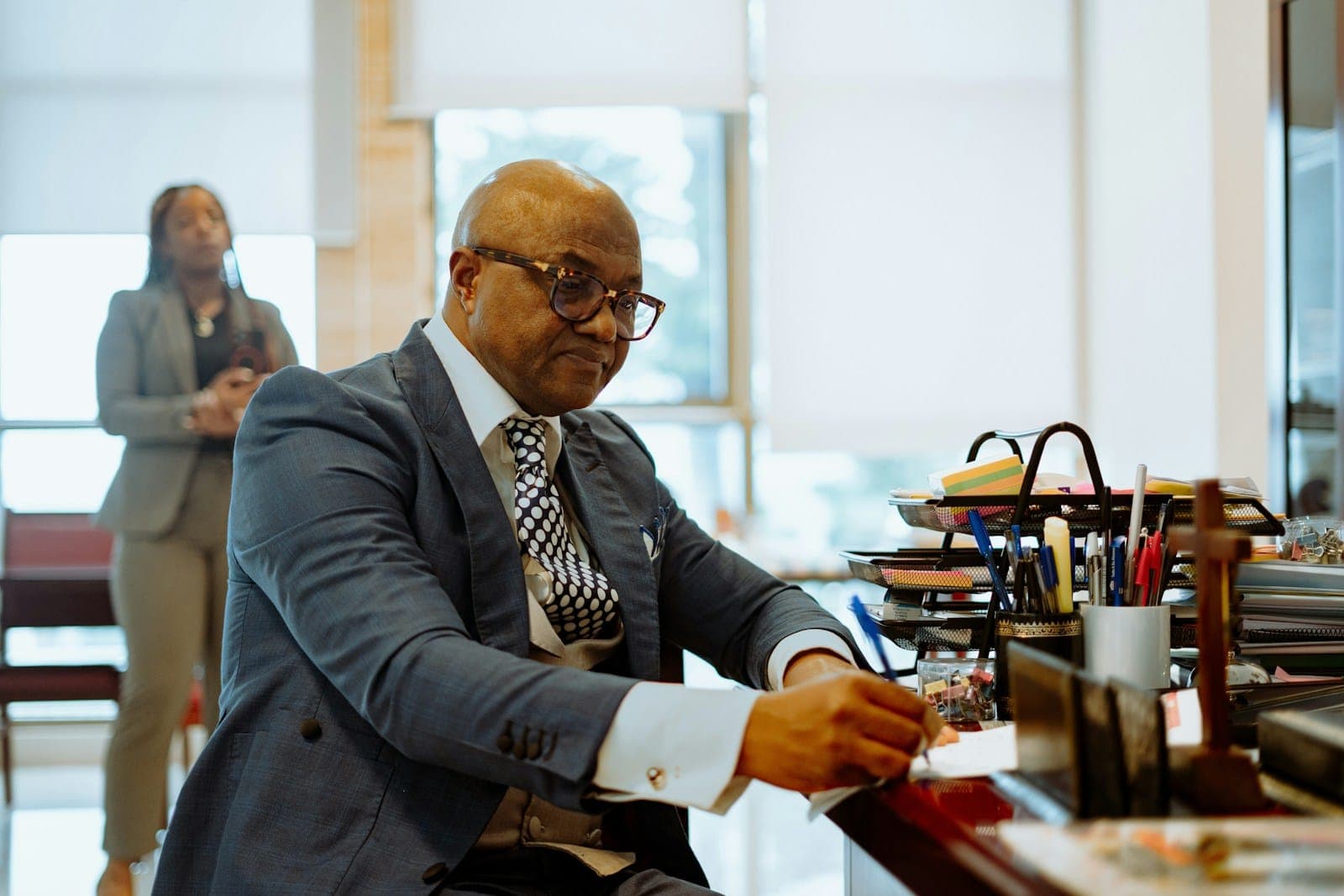 A man in a grey suit and glasses works through papers at his desk while a colleague stands by the window behind him