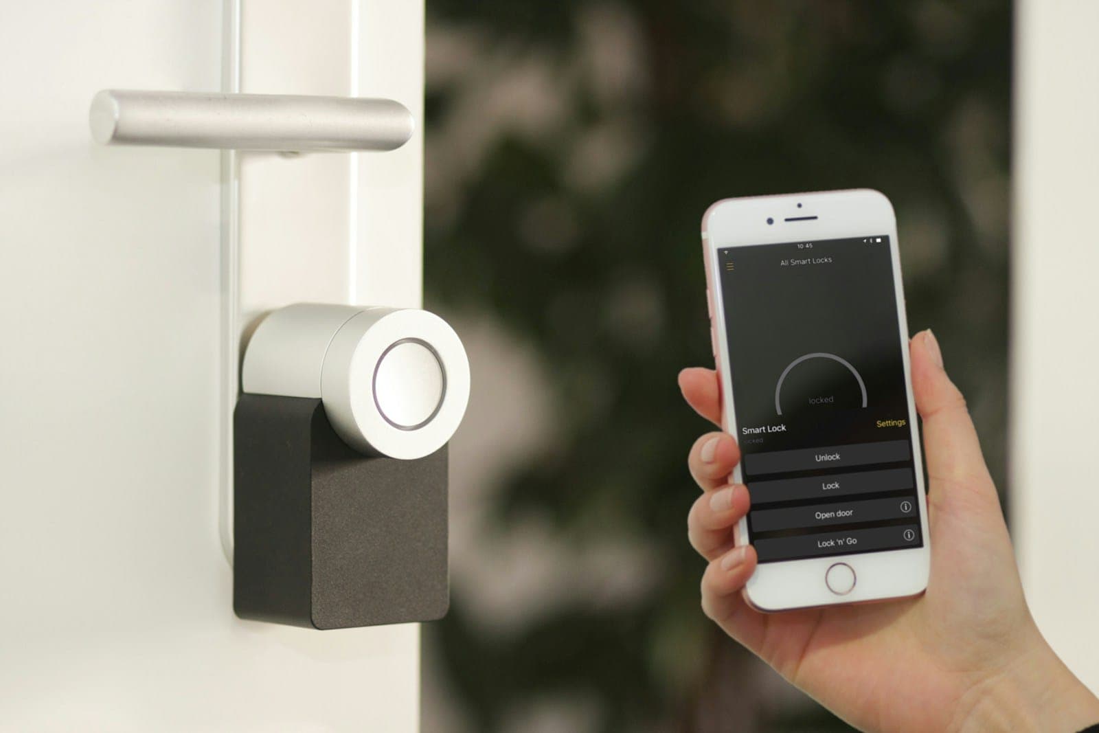 A hand holds a smartphone showing a lock app next to a smart lock fitted to a white door